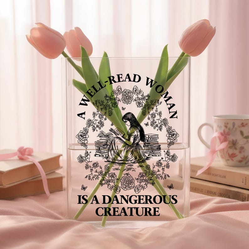 May include: A clear rectangular acrylic vase featuring a black graphic print of a person reading among stacks of books and floral wreaths. The text reads A WELL-READ WOMAN IS A DANGEROUS CREATURE. Three pink tulips are placed inside the vase.