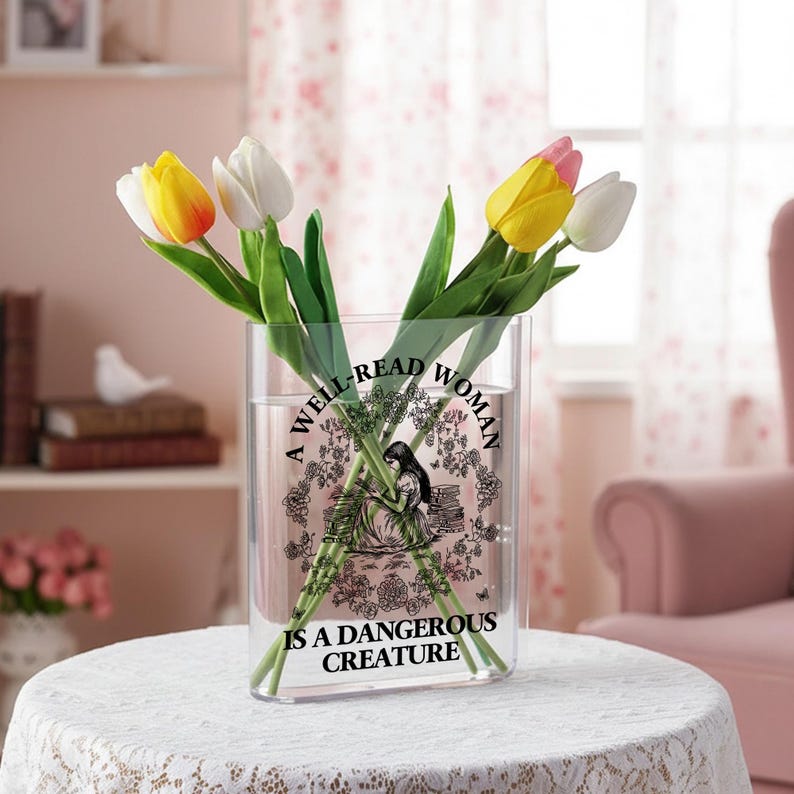 May include: A clear rectangular acrylic vase featuring a black graphic illustration of a person sitting among books, surrounded by a floral wreath. The text reads A WELL-READ WOMAN IS A DANGEROUS CREATURE. The vase holds yellow, white, and pink tulips.