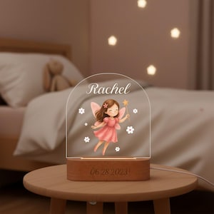 May include: A personalized night light with a wooden base and a clear acrylic top. The acrylic features a cartoon fairy in a pink dress, the name "Rachel", and the date "06.28.2023". The light is on a small wooden table.