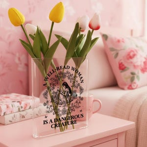 May include: A clear, rectangular acrylic vase featuring a black graphic illustration of a person reading among flowers and books. The text reads, A WELL-READ WOMAN IS A DANGEROUS CREATURE. The vase contains four artificial tulips in yellow and white.