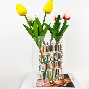 Pode incluir: Um vaso retangular transparente com um design de estante cheio de tulipas amarelas e brancas. O vaso tem uma base curva e est&aacute; sobre um livro. O design da estante apresenta livros coloridos e detalhes florais.