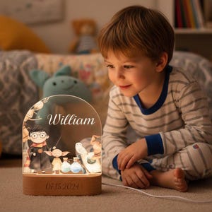 Peut inclure: Une veilleuse personnalisée avec une base en bois et un dessus transparent en forme d'arche. Le dessus présente une illustration de personnages de dessins animés, le nom "William" et la date "07.15.2024". La lumière est branchée et illumine la pièce.