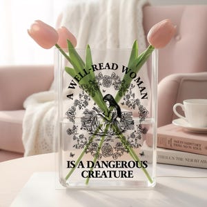 May include: A clear rectangular glass vase containing three light pink tulips. The front of the vase features a black graphic print of a person reading among books and flowers, with the text A WELL-READ WOMAN IS A DANGEROUS CREATURE.