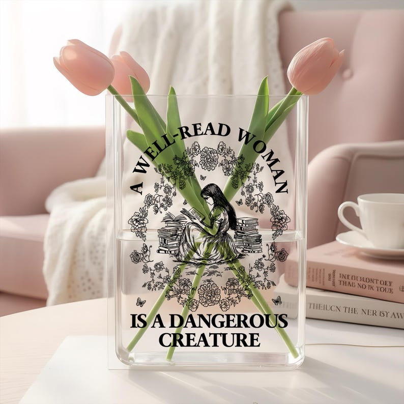 May include: A clear rectangular glass vase containing three light pink tulips. The front of the vase features a black graphic print of a person reading among books and flowers, with the text A WELL-READ WOMAN IS A DANGEROUS CREATURE.
