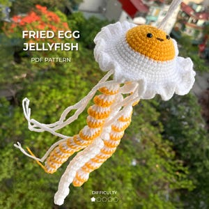 Puede incluir: Una medusa de ganchillo con forma de huevo frito. Tiene una yema amarilla, un borde blanco y rizado, y tentáculos a rayas blancas y amarillas. El texto "FRIED EGG JELLYFISH PDF PATTERN" está incluido en la imagen.