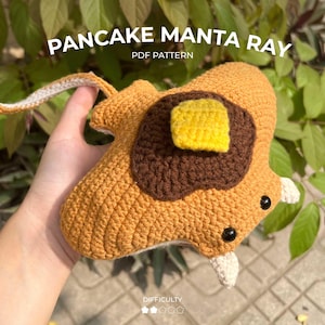 May include: A crocheted manta ray amigurumi toy with a brown pancake design and a yellow crocheted butter topping. The toy is held in a hand and has a white tail.