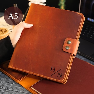 May include: A tan leather A5 notebook cover featuring a snap closure with two metal buttons and the engraved initials H/N on the front cover. The notebook cover has visible stitching along the edges and a smooth, matte finish on the exterior surface.