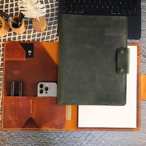 May include: A brown leather portfolio with a green leather cover, a phone pocket, and a pen holder. The portfolio has a white paper insert and a flap with a snap closure.