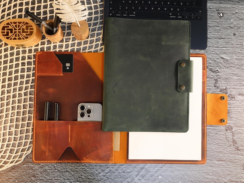 May include: A brown leather portfolio with a green leather cover, a phone pocket, and a pen holder. The portfolio has a white paper insert and a flap with a snap closure.