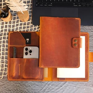 May include: A brown leather notebook with a built-in phone pocket and pen holder. The notebook has a flap that closes with a snap. The notebook is open and has a blank page inside.