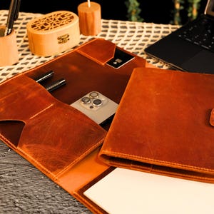 May include: A brown leather portfolio with a built-in phone pocket and pen holders. The portfolio has a flap that closes with a snap and a separate notebook with a strap.