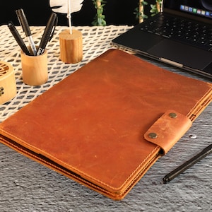 Op de afbeelding: Een bruin leren notitieboek met een riemsluiting en messingkleurige klinknagels. Het notitieboek is open en toont de pagina's. Een zwarte pen ligt op het oppervlak naast het notitieboek. Andere items zijn pennen in een houten houder en een laptop.