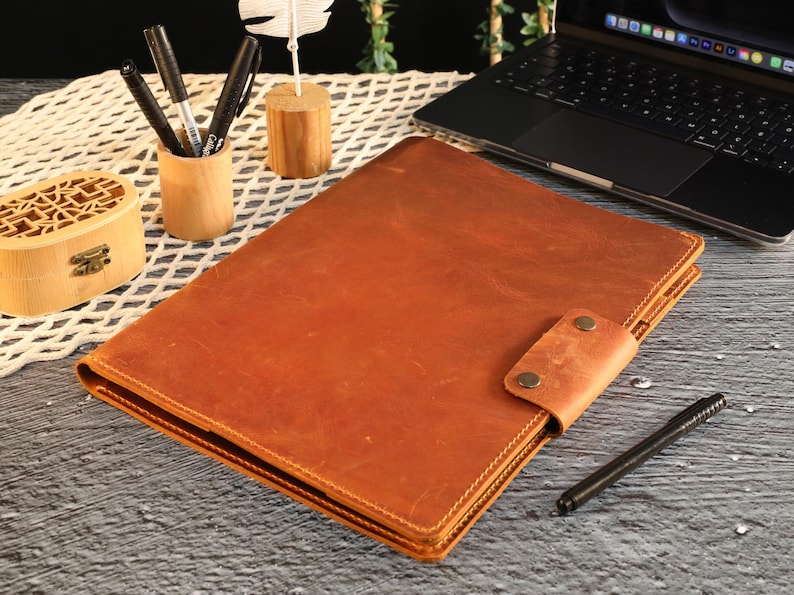 May include: A brown leather folder with a flap closure and a metal clasp. The folder is lying on a wooden surface.