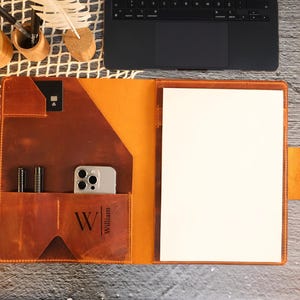 Può includere: Portfolio in pelle aperto nei toni del marrone e dell'arancione, con un foglio di carta bianco all'interno. Dispone di tasche per penne, un telefono e una carta. Il nome "William" è stampato sul davanti. Un laptop e accessori da scrivania sono sullo sfondo.