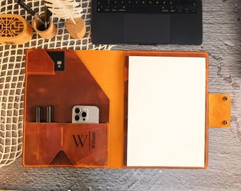 May include: Open leather portfolio in shades of brown and orange, with a white sheet of paper inside. Features pockets for pens, a phone, and a card. The name "William" is printed on the front. A laptop and desk accessories are in the background.