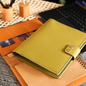 May include: A chartreuse leather journal with a snap closure sits atop an open orange leather notebook. A laptop and desk accessories are in the background. The journal is approximately 8 inches long and 6 inches wide.