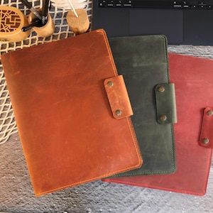 May include: Three leather folders in different colors: brown, green, and red. Each folder has a flap with a metal closure.