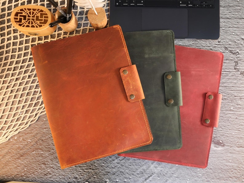 May include: Three leather folders in different colors: brown, green, and red. Each folder has a flap with a metal closure.