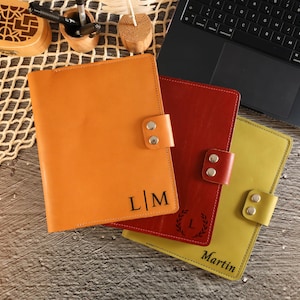 May include: Three leather journals in orange, red, and yellow, each with a snap closure. The orange journal has the initials "L|M", the red journal has a laurel wreath with the letter "L", and the yellow journal has the name "Martin".