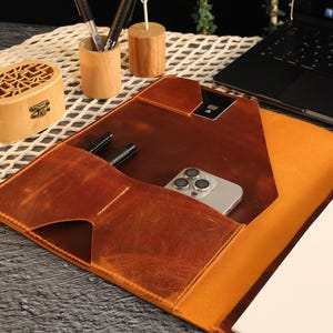 May include: A brown leather portfolio with a flap closure and a built-in pocket for a phone or other small items. The portfolio has a notepad and pen holder and is open to reveal a blank page.