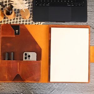 May include: A brown leather portfolio with a flap closure and a built-in pocket for a phone or other device. The portfolio has a white paper insert and a brown leather strap with a metal buckle.