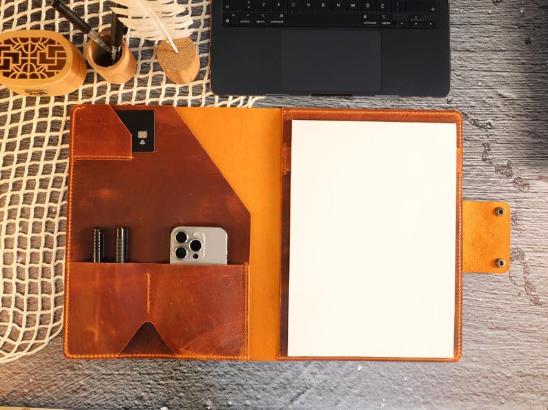 May include: A brown leather portfolio with a flap closure and a built-in pocket for a phone or other device. The portfolio has a white paper insert and a brown leather strap with a metal buckle.