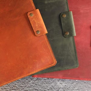 May include: Three leather folders in different colors: orange, green, and red. Each folder has a flap with a metal closure.