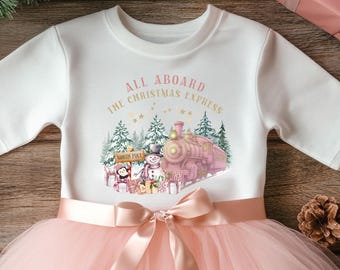May include: White long-sleeved top with a pink tutu skirt. The top features a graphic of a pink train, snowman, and penguin with the text "All Aboard The Christmas Express". A pink bow adorns the waist.