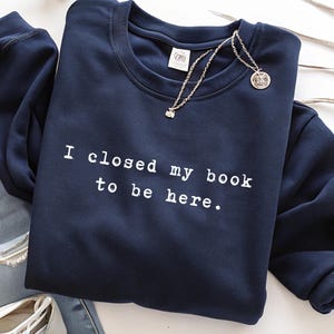Op de afbeelding: Marineblauwe sweatshirt met de witte tekst "I closed my book to be here". Een zilveren ketting met een kleine hanger ligt op de trui. De sweatshirt heeft een ronde hals.