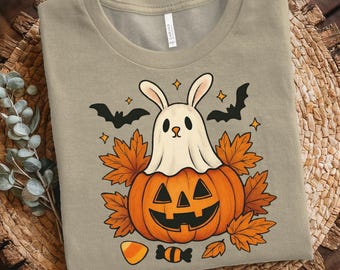Puede incluir: Una camiseta color beige con un diseño de Halloween. El diseño incluye una calabaza con un conejo fantasmal, hojas de otoño, murciélagos, estrellas y caramelos. La camiseta está hecha de un material suave.
