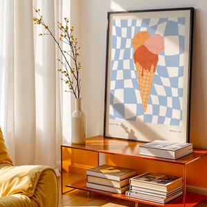 May include: A framed art print featuring an ice cream cone with three scoops in shades of orange and pink, set against a blue and white checkered background. The print is displayed on an orange cabinet with books and a vase of flowers.