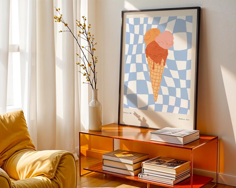 May include: A framed art print featuring an ice cream cone with three scoops in shades of orange and pink, set against a blue and white checkered background. The print is displayed on an orange cabinet with books and a vase of flowers.