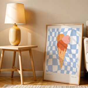 May include: A framed art print featuring a colorful ice cream cone with orange, pink, and red scoops on a blue and white checkered background. The print is displayed in a light wood frame, leaning against a wall next to a wooden side table and lamp.