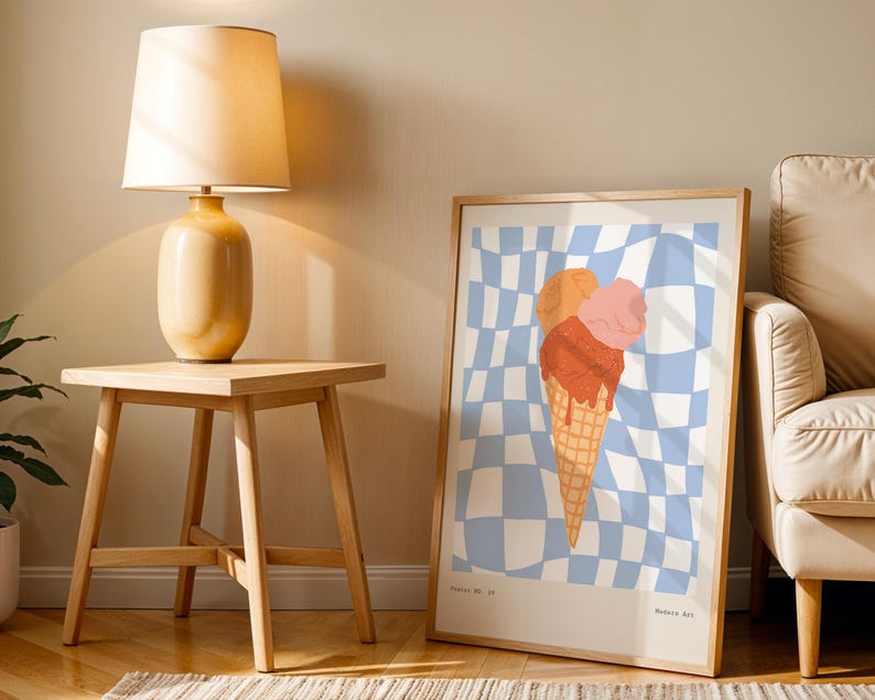 May include: A framed art print featuring a colorful ice cream cone with orange, pink, and red scoops on a blue and white checkered background. The print is displayed in a light wood frame, leaning against a wall next to a wooden side table and lamp.