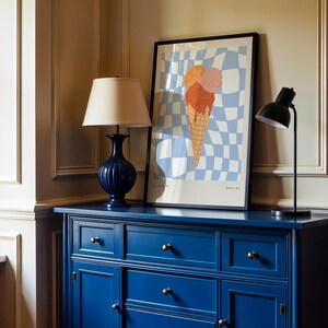 May include: A navy blue cabinet with gold knobs supports a framed ice cream cone art print and two lamps. The art features a checkered background with a large ice cream cone. A dark blue lamp with a beige lampshade and a black desk lamp are on the cabinet.