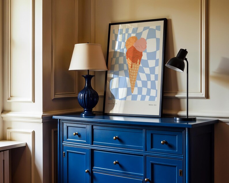 May include: A navy blue cabinet with gold knobs supports a framed ice cream cone art print and two lamps. The art features a checkered background with a large ice cream cone. A dark blue lamp with a beige lampshade and a black desk lamp are on the cabinet.