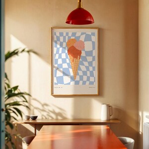 May include: A framed art print featuring an ice cream cone with three scoops in shades of orange and pink. The background is a blue and white checkered pattern. The print is displayed on a beige wall in a room with a red light fixture.