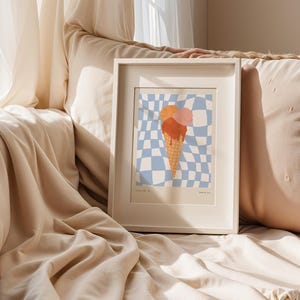 May include: A framed art print featuring an ice cream cone with three scoops in shades of orange and pink. The cone is set against a blue and white checkered background. The frame is white.