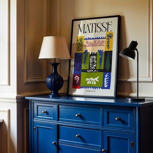 May include: A navy blue cabinet with brass knobs supports a framed "MATISSE" art print and two lamps. One lamp is navy blue with a cream shade, and the other is a black desk lamp. The artwork features colorful abstract shapes.