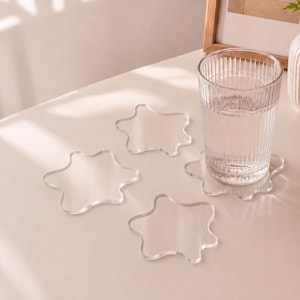 May include: A set of three clear, star-shaped acrylic coasters with smooth, rounded edges. One coaster supports a tall, ribbed glass filled with clear liquid. The coasters are transparent and feature a simple, fluid, five-pointed star design.