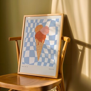 May include: A framed art print featuring an ice cream cone with three scoops in shades of orange and pink. The background is a blue and white checkered pattern. The print is displayed on a wooden chair.