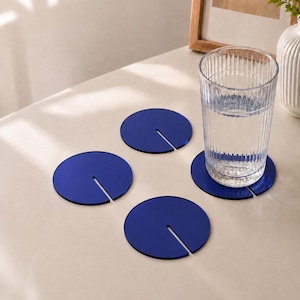 Può includere: Un set di quattro sottobicchieri circolari blu intenso, ciascuno caratterizzato da una fessura dritta che si estende dal bordo al centro. Un sottobicchiere sostiene un bicchiere trasparente a coste verticali pieno d'acqua, mentre altri tre sono appoggiati in piano.