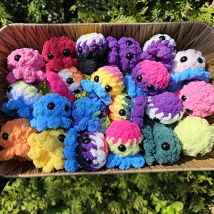 以下が含まれることがあります： カラフルな手作りの編みぐるみのタコが入った柳のバスケット。タコはピンク、赤、青、黄、紫、緑など様々な色があります。各タコは黒いボタンの目とユニークな色の組み合わせを持っています。