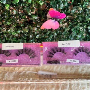 K&ouml;nnte beinhalten: Zwei transparente Plastikboxen mit rosa Deckeln, die falsche Wimpern enthalten. Die Boxen sind mit "Obsession" und "Stay Pretty" beschriftet, mit den entsprechenden Wimpernstilnummern. Jede Box enth&auml;lt einen kleinen schwarzen Wimpernapplikator-Pinsel.