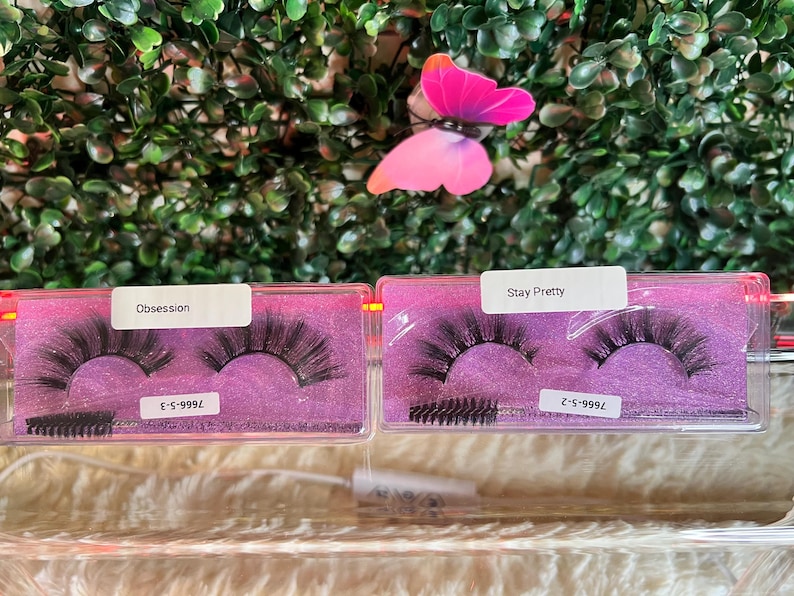K&ouml;nnte beinhalten: Zwei transparente Plastikboxen mit rosa Deckeln, die falsche Wimpern enthalten. Die Boxen sind mit "Obsession" und "Stay Pretty" beschriftet, mit den entsprechenden Wimpernstilnummern. Jede Box enth&auml;lt einen kleinen schwarzen Wimpernapplikator-Pinsel.