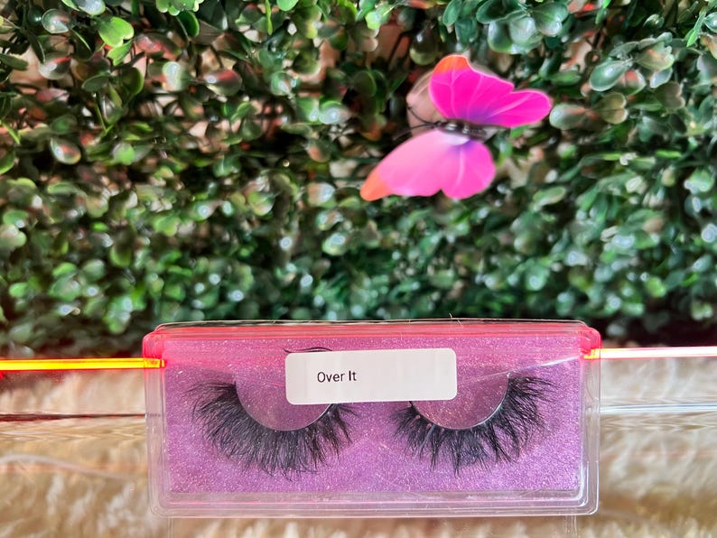 K&ouml;nnte beinhalten: Nahaufnahme eines rosa und durchsichtigen Plastikbeh&auml;lters mit zwei Paaren k&uuml;nstlicher Wimpern. Der Beh&auml;lter tr&auml;gt ein Etikett mit der Aufschrift "Over It".