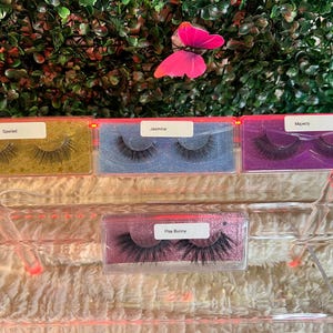 K&ouml;nnte beinhalten: Vier transparente Plastikbeh&auml;lter mit verschiedenen Hintergrundfarben und schwarzen falschen Wimpern. Die Beh&auml;lter sind mit "Spoiled", "Jasmine", "Majesty" und "Play Bunny" beschriftet.