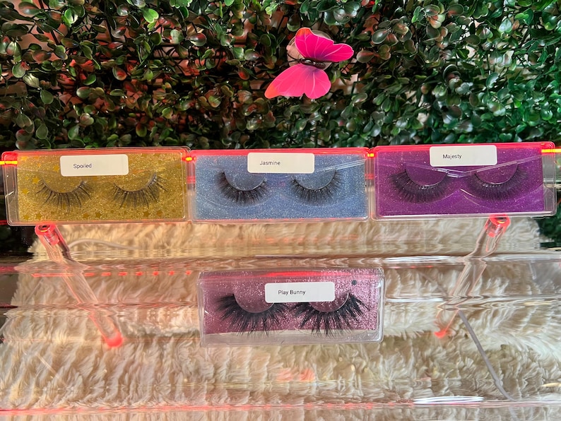 K&ouml;nnte beinhalten: Vier transparente Plastikbeh&auml;lter mit verschiedenen Hintergrundfarben und schwarzen falschen Wimpern. Die Beh&auml;lter sind mit "Spoiled", "Jasmine", "Majesty" und "Play Bunny" beschriftet.