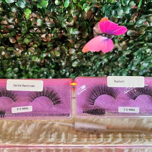 K&ouml;nnte beinhalten: Zwei rosa Boxen mit durchsichtigen Deckeln, die falsche Wimpern enthalten. Die Boxen sind mit "Gentle Reminder" und "Radiant" beschriftet. Die Wimpern sind schwarz und haben ein nat&uuml;rliches Aussehen. Die Boxen stehen auf einer wei&szlig;en Oberfl&auml;che.