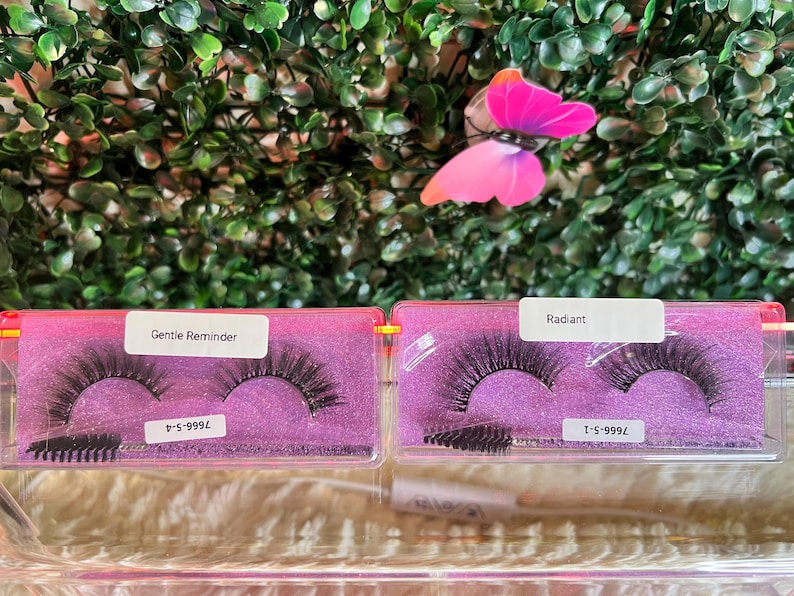 K&ouml;nnte beinhalten: Zwei rosa Boxen mit durchsichtigen Deckeln, die falsche Wimpern enthalten. Die Boxen sind mit "Gentle Reminder" und "Radiant" beschriftet. Die Wimpern sind schwarz und haben ein nat&uuml;rliches Aussehen. Die Boxen stehen auf einer wei&szlig;en Oberfl&auml;che.